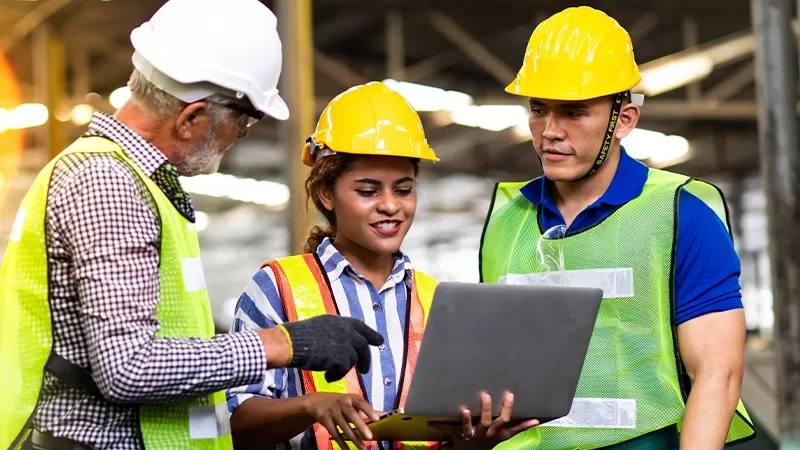 Three people in safety vests and hard hats studying a laptop screen.